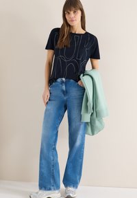 Femme portant un t-shirt graphique bleu marine et un jean bleu à jambes larges, tenant un sweat-shirt vert menthe plié contre un mur beige.