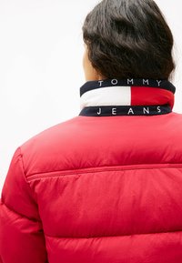 Rød pufferjakke med quiltet tekstur, med en krave i navy og hvid med "TOMMY JEANS" branding. Baghånden viser syningerne i detalje.