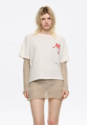 T-shirt in cotone beige con una vestibilità ampia, maniche corte, caratterizzato da un design stampato rosso sul davanti. Indossato sopra un abito in maglia beige trasparente e testurizzato.