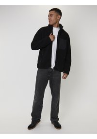 Uomo che indossa un giubbotto in pile nero sopra una maglietta bianca, jeans grigio scuro e sneakers nere, in piedi contro uno sfondo chiaro e uniforme.