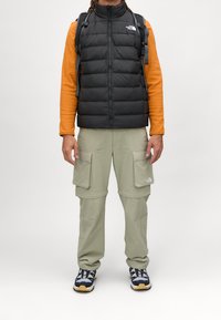 Czarna pikowana kamizelka na pomarańczowej bluzie z długim rękawem, w połączeniu z jasnoskalnymi spodniami typu cargo i trampkami z niebieskimi i białymi akcentami.