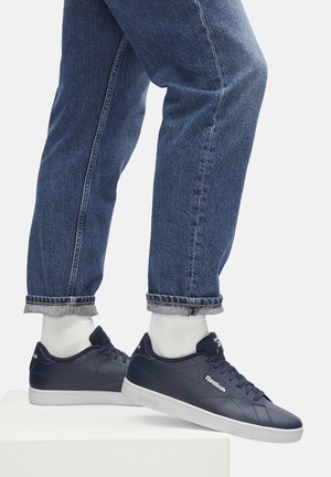 Persoană îmbrăcată cu blugi albaștri cu mansete, șosete albe și adidași Reebok bleumarin, stând pe o platformă albă, pe un fundal alb.