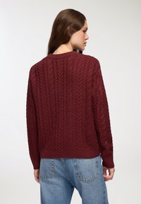 Maglione lavorato a maglia borgogna con motivo a treccia testurizzata, colletto a girocollo a coste e orlo aderente. Indossato con jeans a vita alta azzurri.