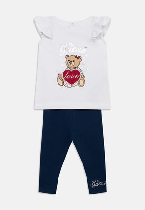 Wit kortermig topje met ruches aan de mouwen, afbeelding van een teddybeer die een rood hart vasthoudt. Navy blauwe legging met logodetail.