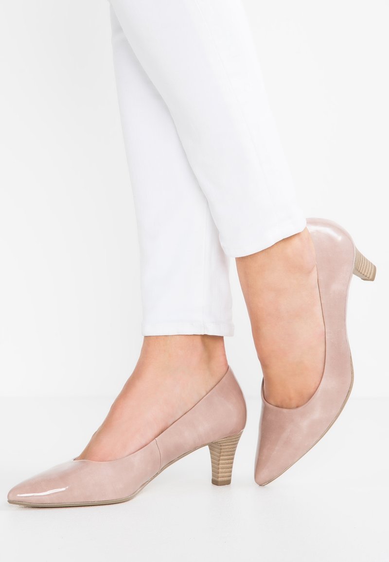 Femme portant un pantalon blanc et des escarpins à talons moyens en cuir verni rose clair, un pied légèrement surélevé derrière l'autre.