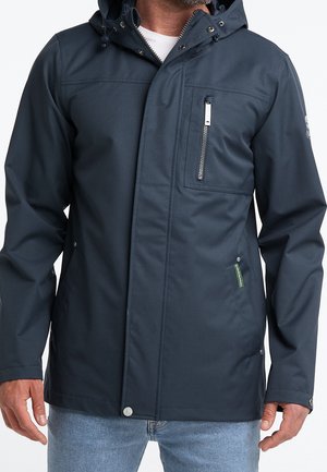 Homme portant une veste à capuche bleu marine avec une poche zippée sur la poitrine et des poches latérales à boutons-pression, associée à un jean bleu clair.