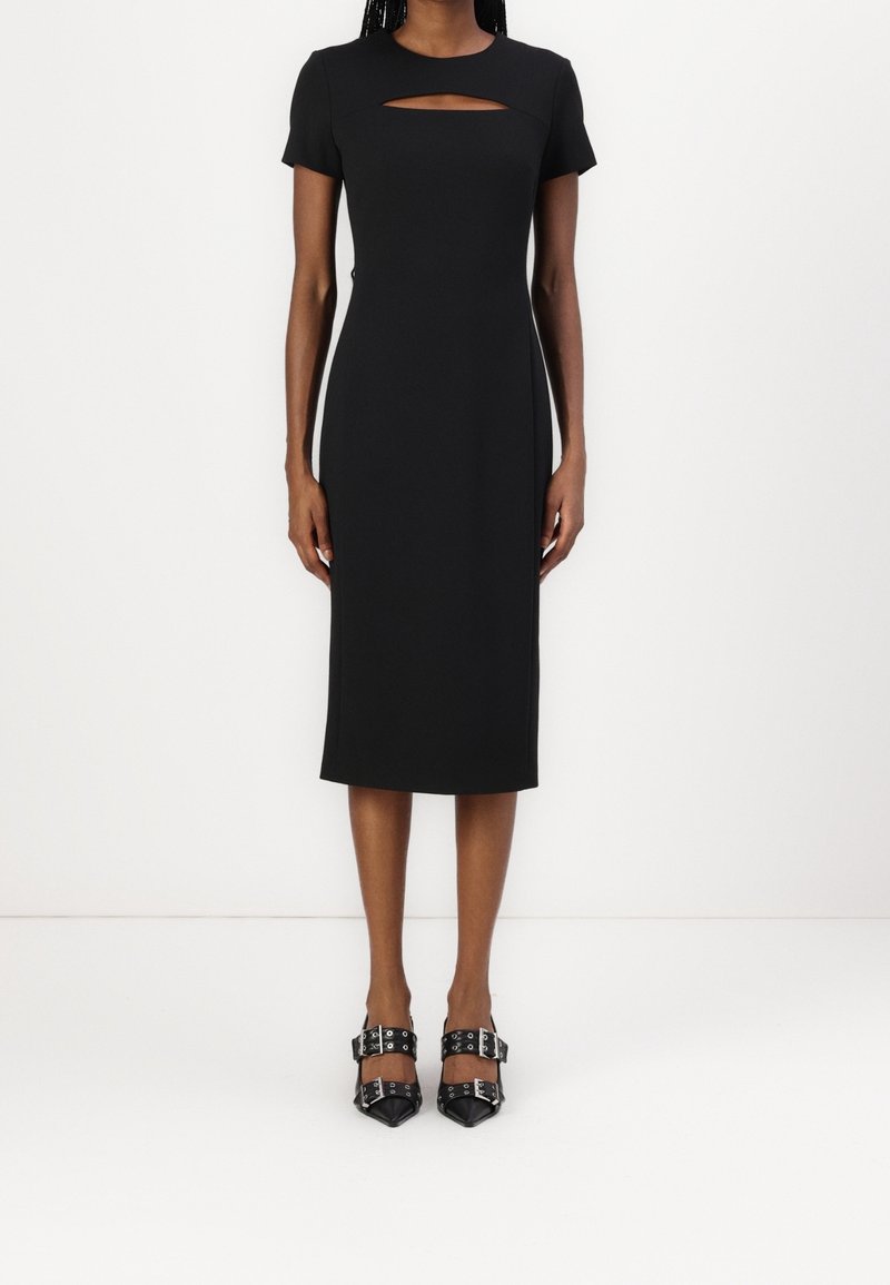 Femme portant une robe midi noire ajustée avec manches courtes et une découpe horizontale sur la poitrine, associée à des chaussures noires pointues à brides bouclées.