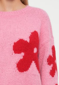 Pullover de malha rosa suave com grandes padrões de flores vermelhas no peito e nas mangas, apresentando um decote redondo e tecido texturizado.