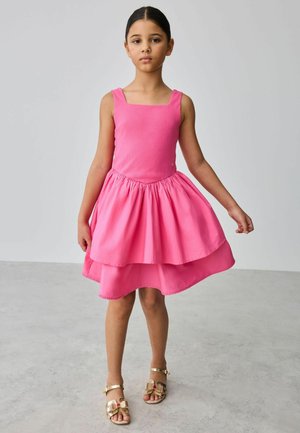 Mladé dievča stojí a nosí ružové vrstvené šaty bez rukávov a zlaté sandále na svetlom neutrálnom pozadí.