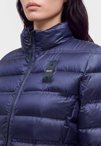 Femme portant une veste matelassée bleu marine brillante avec un col montant et un petit patch logo rectangulaire sur la poitrine.