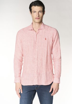 Uomo che indossa una camicia a maniche lunghe a righe rosa e bianche con logo ricamato, abbinata a jeans blu, in piedi su uno sfondo neutro.