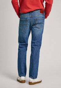 Jeans de denim azul con un corte entallado, cinco bolsillos y detalles de costura sutiles. Llevados con zapatillas blancas y una sudadera roja.