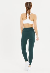 Leggings de tiro alto en verde oscuro con una textura suave, que cuentan con bolsillos laterales y un diseño ajustado, combinados con un sujetador deportivo blanco.