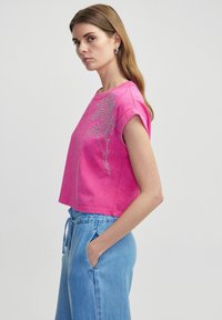 Roze cropped t-shirt van zachte katoen, met een bloemenontwerp in rhinestones op de linker schouder. Draag het met lichtblauwe shorts met een koord.