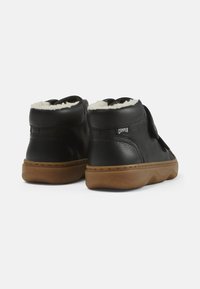 Zapatos de caña alta de cuero negro con un suave forro blanco, cierre de velcro y suela de goma marrón texturizada con un diseño ondulado.