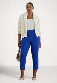 Nő fehér kötött kardigánt és fehér ujjatlan felsőt visel, élénk kék, rövidített nadrágot, bézs balerinacipőt, bézs kézitáskát tart, semleges háttér előtt áll.