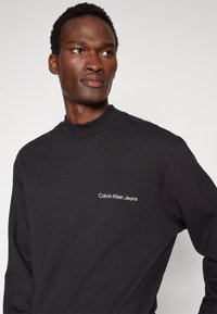Felpa nera con collo alto, caratterizzata da ricamo bianco "Calvin Klein Jeans" sul lato sinistro del petto. Texture liscia e vestibilità morbida.