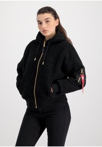 Svart fleece zip-up jacka med huva, guldzip och svarta muddar. Har sidofickor och en liten röd etikett på vänster ärm.