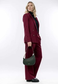 Bordeauxroter maßgeschneiderter Anzug mit einem schwarzen Rollkragenpullover, der eine grüne Lederhandtasche hält. Die Tasche hat eine geschwungene Form und goldene Hardware-Details.