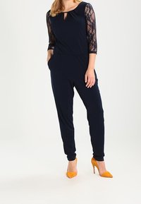 Marinblå jumpsuit med trekvartslånga spetsärmar, med en nyckelhålsringning och sidofickor, kombinerad med gula spetsiga klackskor.