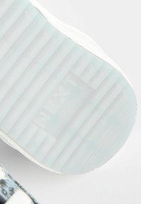 Valge alus, millel on tekstuurilised sooned ja embossed "NEXT" logo. Selge, veidi poolkompleksne materjal võimaldab näha sisemist struktuuri.