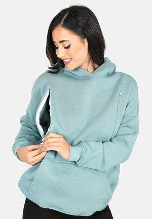 Woman in light blue hoodie unzipping side to reveal inner layer, smiling and looking down, wearing black pants.