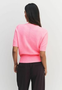 Kratka majica s kratkimi rokavi v pastelno rožnati barvi, izdelana iz pletene tkanine, z ribanim robom in podrobnostmi na hrbtu, v kombinaciji s temnimi hlačami.