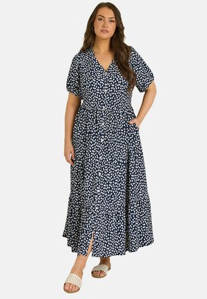 Kobieta ubrana w granatową sukienkę midi w białe kropki, z przodem na guziki, krótkimi bufkami na rękawach, bocznymi kieszeniami i beżowymi sandałami na płaskiej podeszwie.