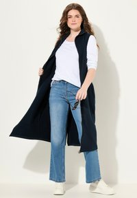 Gilet lungo blu navy con chiusura a zip, indossato sopra una maglietta bianca e jeans azzurri chiari. Il modello tiene degli occhiali da sole e indossa sneakers bianche.