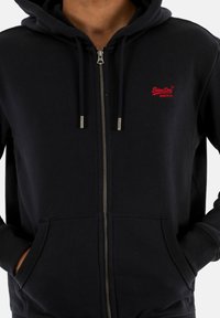 Zwarte rits-gevende hoodie van zacht materiaal, met een rood logo op de linkerkant van de borst, capuchon met trekkkoord en voorzakken.