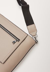 Borsa a spalla in pelle beige con finitura testurizzata, dotata di una tracolla nera, una tasca con cerniera e dettagli in metallo argentato.