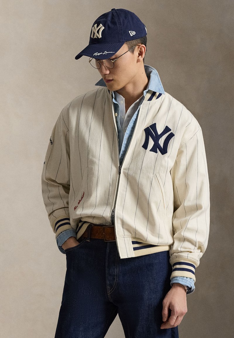 Valge pesapalli jakk mereväe triipude ja NY logoga. Kantud hele sinise kraega särgi peal. Meresinine nokamüts ja tumedad sinised teksad täiendavad rõivastust.