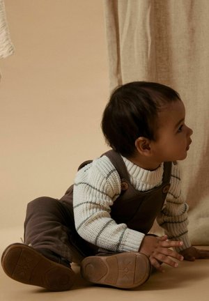 Brune overalls over en lys, stripete genser. Barnet har mørkt hår, et rundt ansikt, og sitter på en beige overflate.