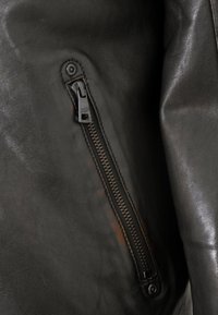 Detail einer schwarzen Lederjacke mit seitlicher Tasche und Reißverschluss, silbernen Beschlägen und glatter Textur.