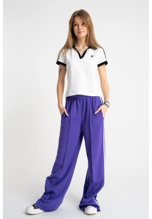 Młoda kobieta z długimi włosami, ubrana w białą koszulę z czarnym obszyciem, szerokie fioletowe spodnie w białe paski oraz białe tenisówki, stoi z rękami w kieszeniach.
