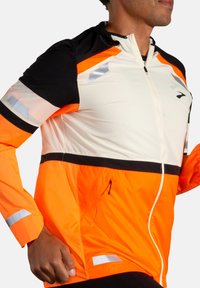 Veste zippée orange et noire avec des accents réfléchissants, manches longues et un ourlet ajustable. Le tissu semble léger et résistant aux intempéries.