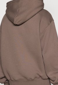 Sweat-shirt à capuche marron avec un tissu texturé et épais. Présente des épaules tombantes et des poignets côtelés, avec un détail de design subtil à l'arrière.