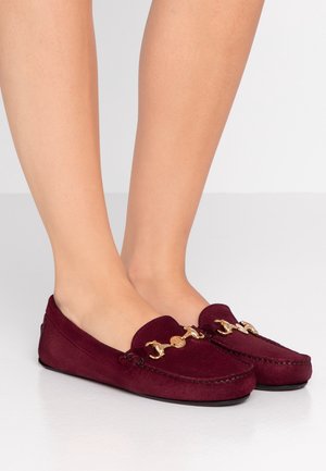 Fødder iført bordeauxrøde mokkasiner i ruskind med guldkædedetalje, sat mod en neutral lys baggrund.