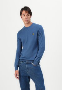 Pull-over en tricot bleu avec un col rond et des poignets et un ourlet côtelés ; arbore un petit logo jaune sur la poitrine. Porté avec un jean en denim foncé.