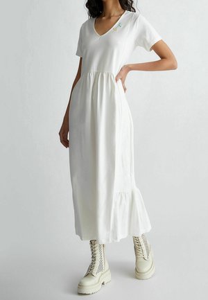 Kvinde iført en hvid maxikjole med korte ærmer og V-hals med flæsekant og cremefarvede snørestøvler i militærstil, hånd på hoften.