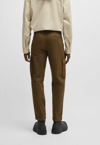 Bruine cargobroek met zijzakken, tapered fit en gestikte details. Draag met zwarte schoenen en een lichtgekleurde lange mouwen shirt.