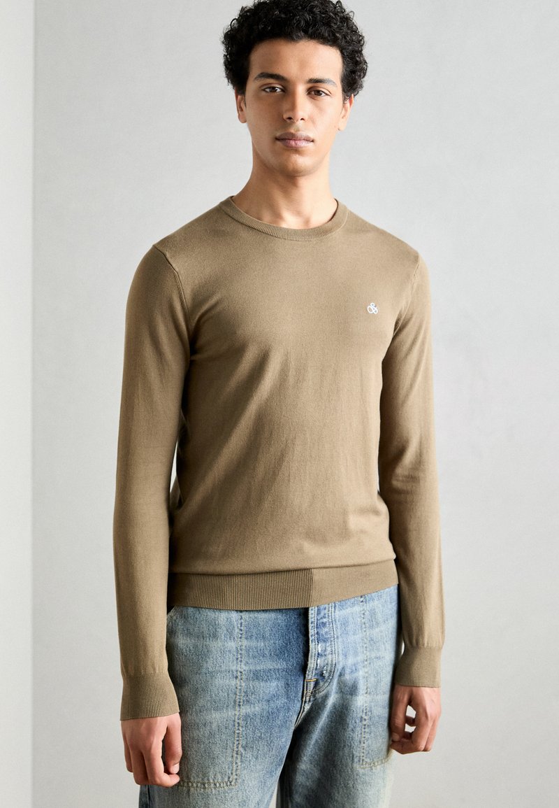 Bruine gebreide trui met lange mouwen, ronde hals en geribbelde zoom; heeft een klein, wit logo op de linkerborst. Draag het met vervaagde blauwe jeans.