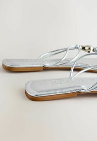 Sandales plates argentées à fines lanières et semelles en bois sur un fond neutre, montrant le profil des orteils et de côté.