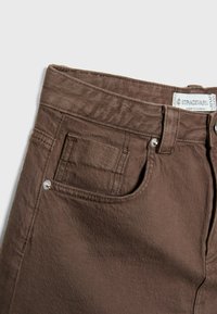 Primo piano di jeans in denim marrone che mostrano la tasca frontale, la tasca porta monetine, la passante per la cintura e l'etichetta Stradivarius cucita all'interno della vita.