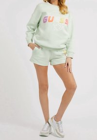 Mujer con sudadera ligera verde y letras coloridas de "GUESS", pantalones cortos a juego y zapatillas blancas, de pie con una mano en el bolsillo.