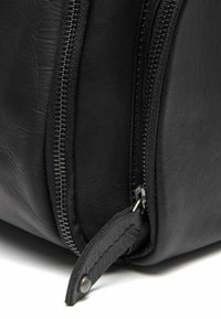 Esquina de una bolsa de cuero negro con cremallera visible, que presenta un material texturizado, herrajes de plata y una pequeña lengüeta de cuero en la cremallera.