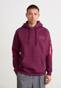 Hoodie bourgogne avec cordons de serrage et poche avant. Dispose d'une poche zippée sur le côté avec un détail de sangle rouge et un patch logo. Tissu doux.