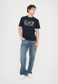 Tamsiai mėlynos spalvos marškiniai su auksiniu EA7 Emporio Armani logotipu, derinami su šviesiai mėlynomis džinsinėmis kelnėmis ir baltais sportbačiais.