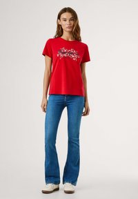 T-shirt rouge à manches courtes avec un motif floral, col rond et coupe décontractée. Associé à un jean évasé bleu clair et des baskets blanches.