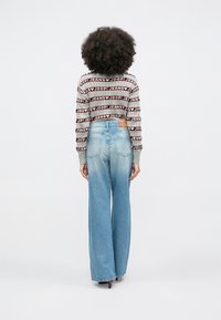 Oseba s kodrastimi lasmi je oblečena v dolg rokav s črtastim vzorcem in napisom "JOOP! JEANS" ter modre kavbojke z visokim pasom in širokimi hlačnicami, obrnjena stran.